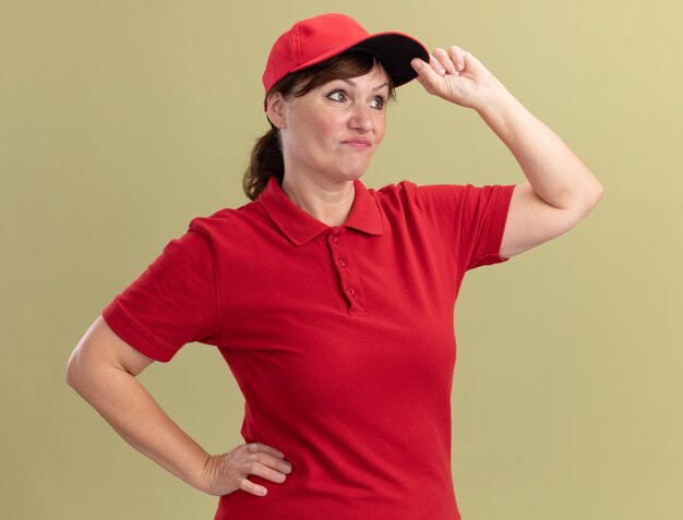 Femme de livraison d'âge moyen en uniforme rouge et cap à côté confus et mécontent de toucher sa casquette debout sur mur vert