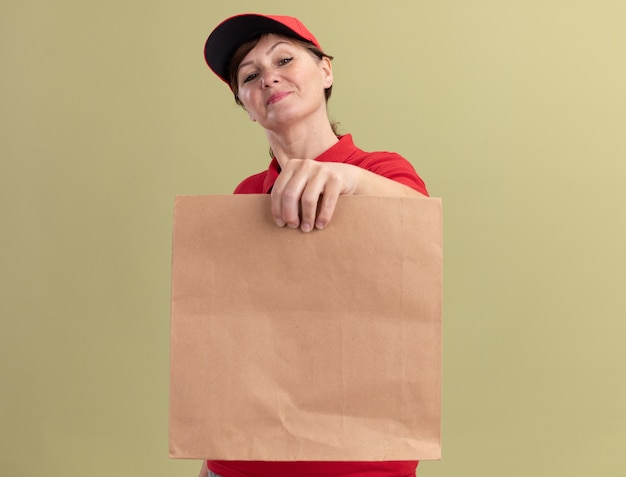 Femme de livraison d'âge moyen en uniforme rouge et cap holding paper package looking at front smiling confiant debout sur mur vert