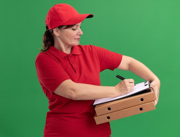 Femme De Livraison D'âge Moyen En Uniforme Rouge Et Cap Tenant Des Boîtes à Pizza Et Presse-papiers Avec Des Pages Vierges écrivant Avec Un Visage Sérieux Debout Sur Un Mur Vert