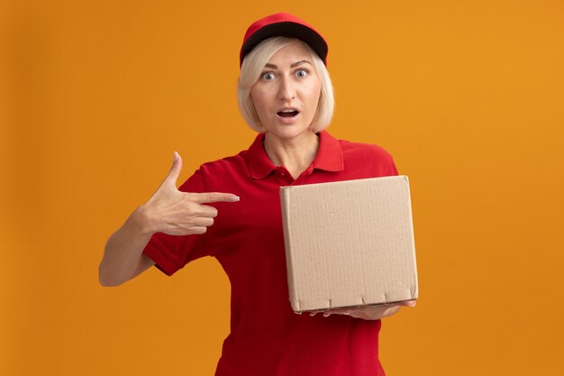 Femme de livraison blonde d'âge moyen impressionnée en uniforme rouge et casquette tenant et pointant sur une boîte en carton regardant à l'avant isolée sur un mur orange avec espace de copie