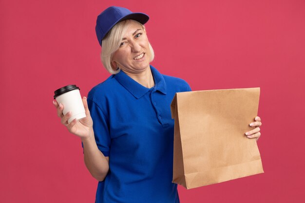 Femme de livraison blonde d'âge moyen mécontente en uniforme bleu et casquette tenant un paquet de papier et une tasse à café en plastique