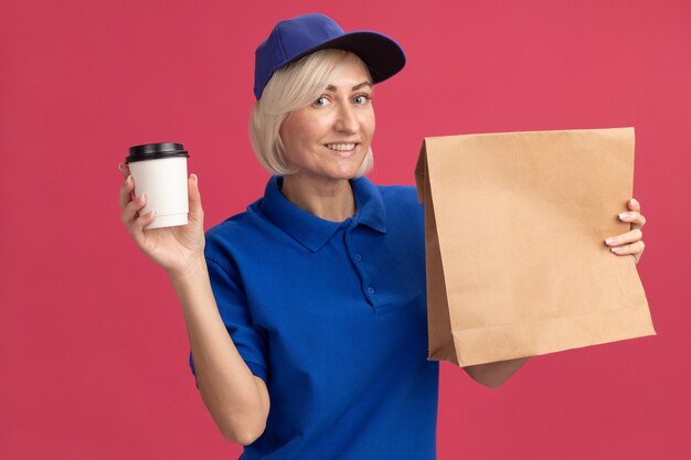 Femme de livraison blonde d'âge moyen souriante en uniforme bleu et casquette tenant un paquet de papier et une tasse à café en plastique