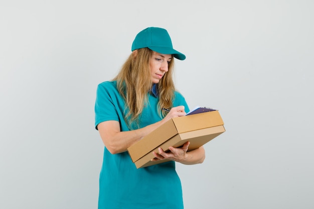 Femme de livraison écrit sur le presse-papiers sur une boîte en carton en t-shirt, casquette et à occupé