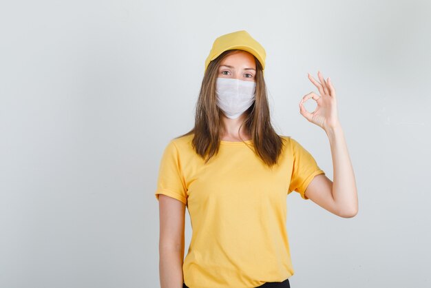 Femme de livraison montrant le geste ok en t-shirt, pantalon, casquette et masque et à la joie.