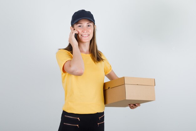 Femme de livraison parlant sur smartphone avec boîte en carton en t-shirt, pantalon et casquette