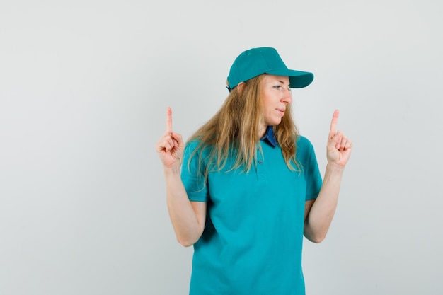 Femme de livraison pointant vers le haut tout en regardant de côté en t-shirt, casquette et à la recherche positive.