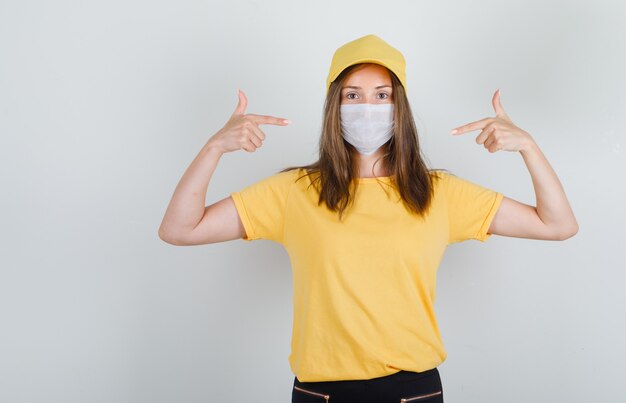 Femme de livraison en t-shirt, pantalon, capuchon pointant du doigt son masque et à la prudence