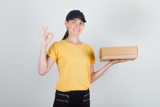 Femme de livraison en t-shirt, pantalon, casquette tenant une boîte en carton avec signe ok et à la satisfaction