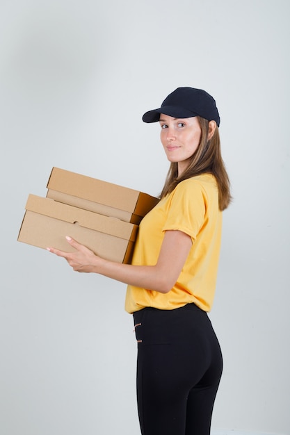 Femme de livraison en t-shirt, pantalon, casquette tenant des boîtes en carton et souriant