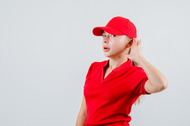 Femme de livraison en t-shirt rouge et casquette en gardant la main derrière l'oreille et à la curiosité