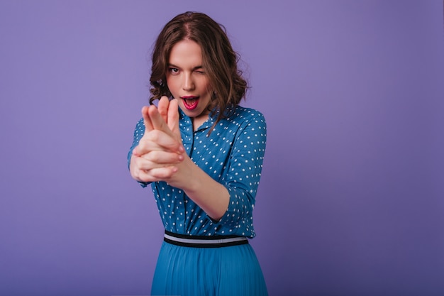 Femme ludique aux cheveux courts en tenue tendance posant sur un mur violet. Modèle féminin de bonne humeur avec des cheveux ondulés s'amusant