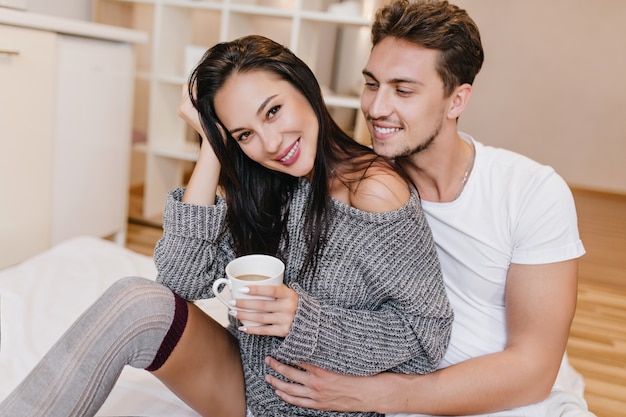 Femme ludique en tenue grise passer la matinée avec son petit ami et profiter d'un café chaud