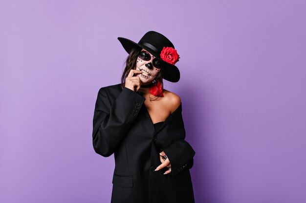 Femme mafieuse pathétique en tenue noire touche ses dents et pose en masque de crâne sur un mur isolé lilas.