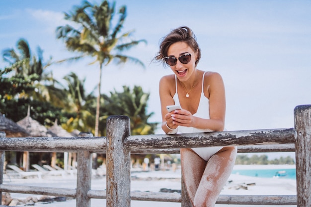 Femme en maillot de bain blanc au bord de l'océan à l'aide d'un téléphone