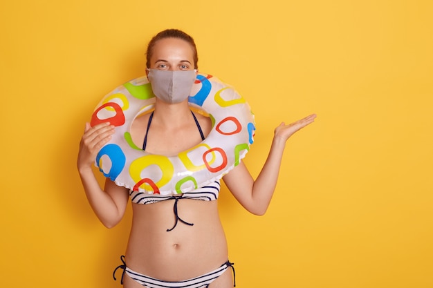 Femme en maillot de bain rayé tenant un anneau en caoutchouc sur son cou, portant un masque hygiénique, repos et vacances d'été avec covid-19. Femelle écartant la paume de côté. Copiez l'espace.