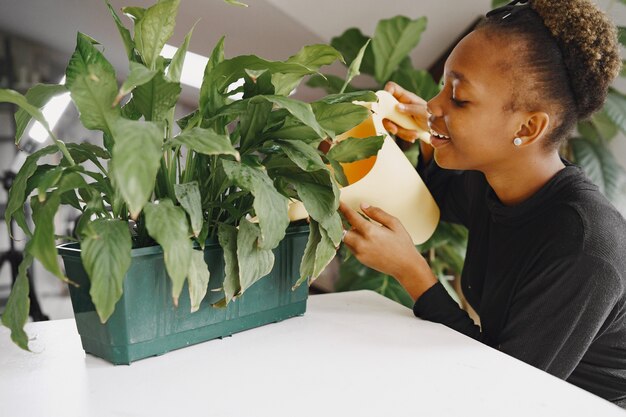 Femme à la maison. Fille dans un pull noir. Femme africaine arrosant la plante. Personne avec pot de fleurs.