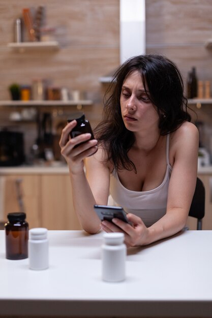 Femme malade souffrant de migraine regardant un récipient contenant des médicaments et un smartphone pour prescription. Personne souffrant de maux de tête tenant une bouteille de médicaments, de médicaments, de remèdes, de médicaments, de traitement