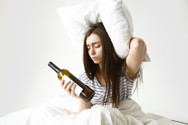 Femme malheureuse a souligné avec les cheveux lâches en désordre, souffrant de maux de tête après la fête, tenant un oreiller et une bouteille vide