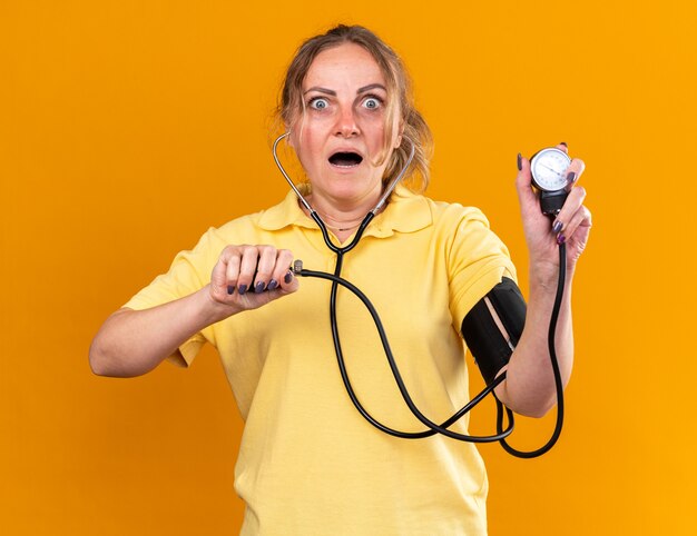 Femme malsaine en chemise jaune ne se sentant pas bien souffrant de la grippe et du froid mesurant sa tension artérielle à l'aide d'un tonomètre l'air inquiète debout sur un mur orange