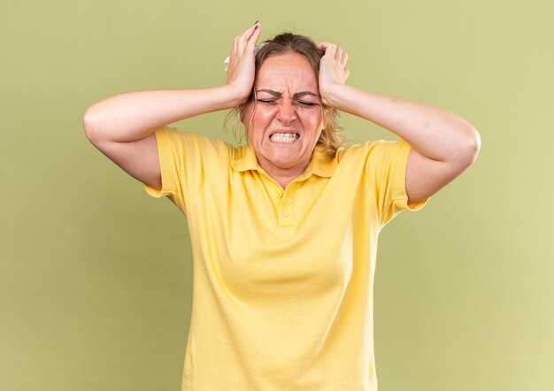 Femme malsaine en chemise jaune se sentant terriblement touchée par la tête souffrant de forts maux de tête ayant la grippe debout sur un mur vert