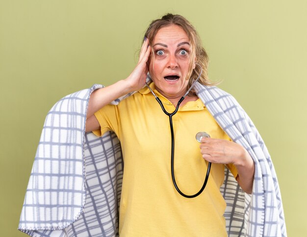 Femme malsaine enveloppée dans une couverture se sentant mal souffrant de la grippe et du froid en écoutant son rythme cardiaque à l'aide d'un stéthoscope l'air inquiet debout sur un mur vert