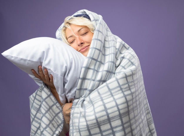 Femme malsaine malade aux cheveux courts dans un chapeau chaud enveloppé dans une couverture tenant un oreiller appuyé sa tête sur un oreiller avec les yeux fermés smilingover fond violet