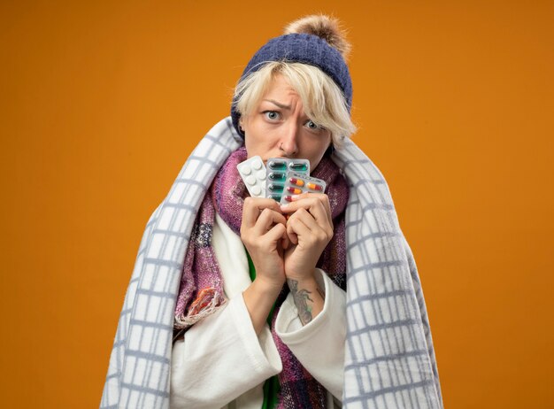 Femme malsaine malade aux cheveux courts dans une écharpe chaude et un chapeau enveloppé dans une couverture tenant des pilules avec une expression triste debout sur un mur orange