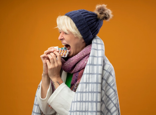 Photo gratuite femme malsaine malade aux cheveux courts dans une écharpe chaude et un chapeau enveloppé dans une couverture tenant des pilules en les mettant tous dans la bouche debout sur un mur orange