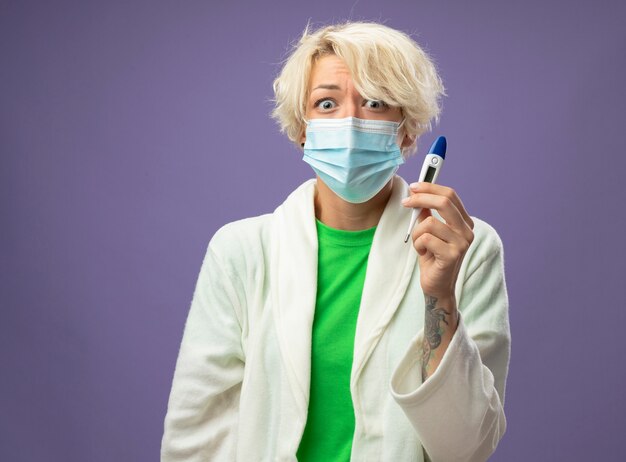 Femme malsaine malade aux cheveux courts dans un masque de protection du visage tenant un thermomètre inquiet sur mur violet