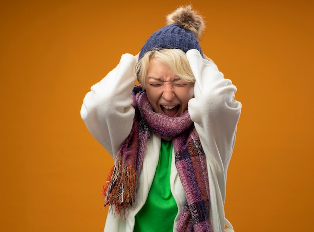 Photo gratuite femme malsaine malade aux cheveux courts en écharpe chaude et chapeau criant et criant avec une expression agressive avec les mains levées debout sur fond orange