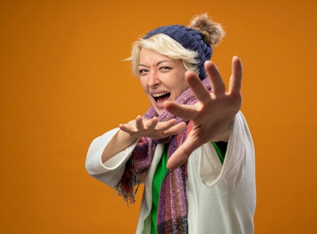 Photo gratuite femme malsaine malade aux cheveux courts en écharpe chaude et chapeau faisant un geste de défense avec les mains effrayées debout sur fond orange