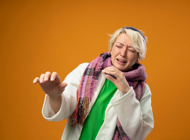 Femme malsaine malade aux cheveux courts en écharpe chaude et chapeau faisant le geste de défense avec les mains avec l'expression de la peur debout sur le mur orange