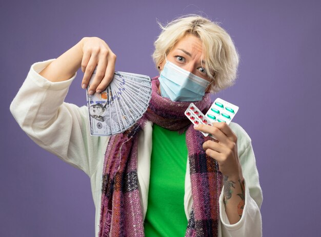 Femme malsaine malade aux cheveux courts en écharpe chaude et masque de protection faciale tenant de l'argent et des ampoules avec des pilules regardant la caméra avec une expression triste debout sur fond violet