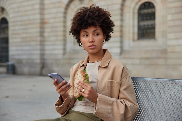 Une Femme Mange Un Délicieux Sandwich Et Utilise Des Repos De Téléphone Portable Après Avoir Traversé La Ville Pose à L'extérieur Sur Un Banc Concentré Loin