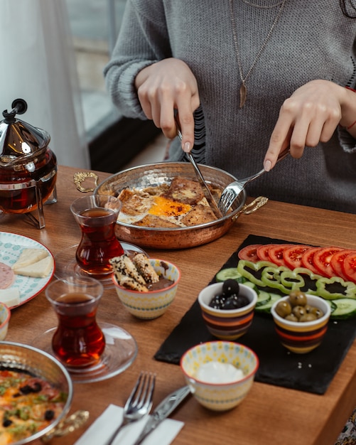 Une femme mange une omelette avec du thé noir et des aliments tout autour.