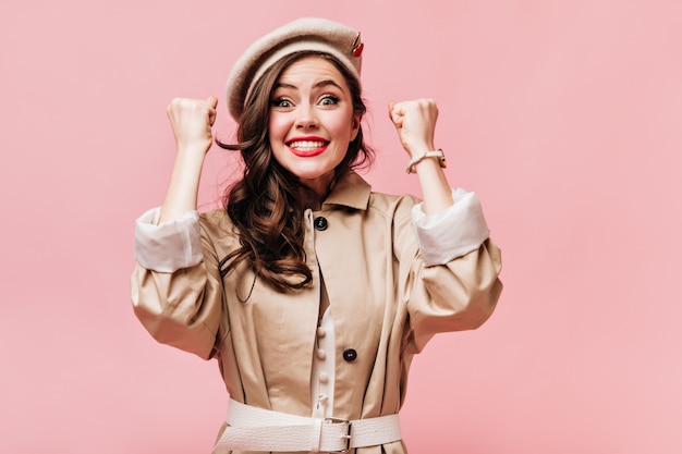 La femme en manteau beige est heureuse de la chance et regarde la caméra avec le sourire sur fond rose.