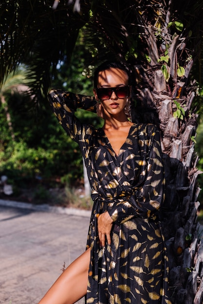 Femme avec maquillage en bronze portant une robe dorée noire et des lunettes de soleil dans un paysage tropical