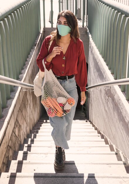 Femme avec masque facial à monter les escaliers tout en tenant des sacs d'épicerie