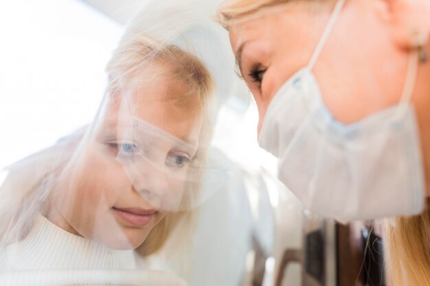 Femme avec masque médical en quarantaine derrière la fenêtre avec fille