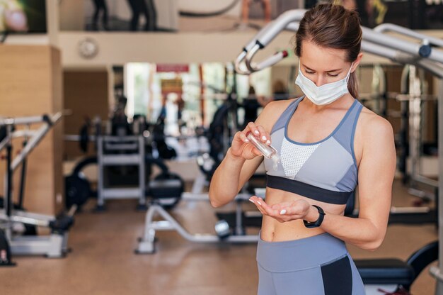 Femme avec masque médical à la salle de sport à l'aide de désinfectant pour les mains