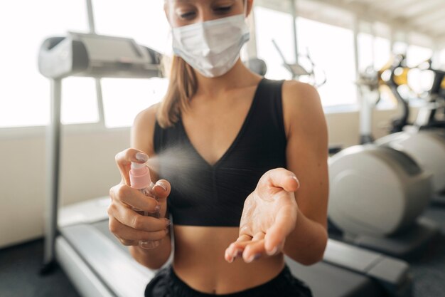 Femme avec masque médical à la salle de sport à l'aide de désinfectant pour les mains