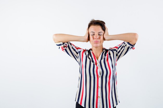 Femme mature en appuyant sur les mains sur les oreilles, fermant les yeux en chemise rayée et à la vue calme, de face.