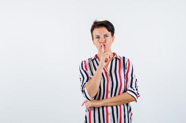 Femme mature en chemise rayée montrant le geste de silence et à la confiance, vue de face.