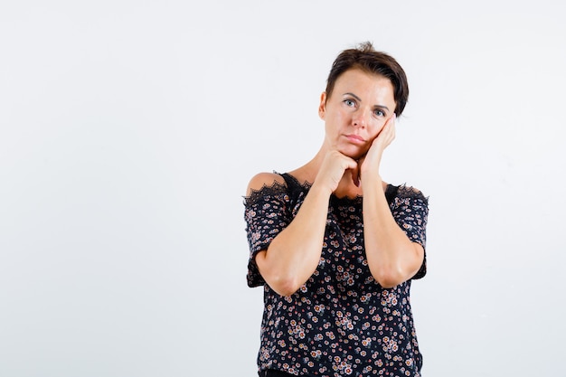 Femme mature en chemisier floral, jupe noire penchée sur la joue sur la paume, étayer le menton sur place, penser à quelque chose et à la recherche pensive, vue de face.