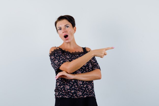 Femme mature en chemisier pointant vers le côté droit et à la perplexité, vue de face.