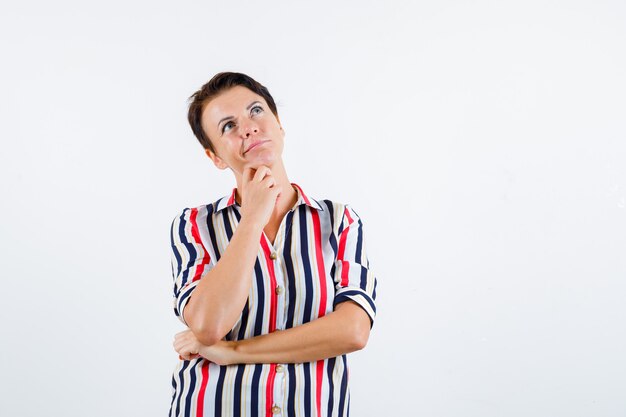 Femme mature en chemisier rayé s'appuyant le menton sur la paume, en pensant à quelque chose et à la recherche pensive, vue de face.