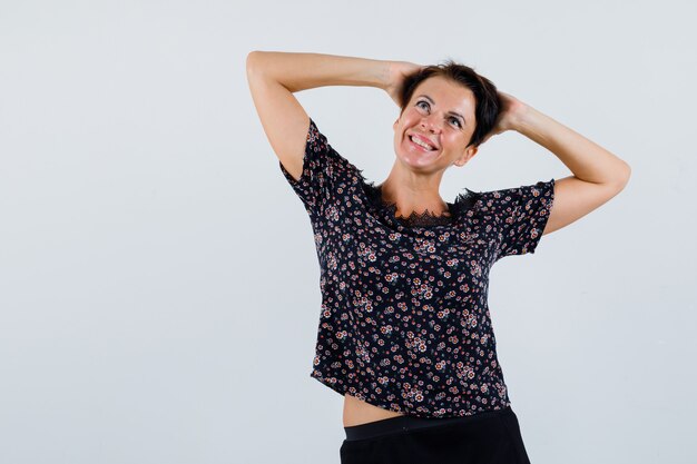 Femme mature en chemisier, tenant les mains derrière la tête tout en levant et à la vue de face, joyeuse.