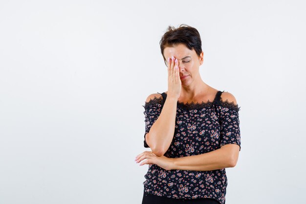 Femme mature couvrant un œil avec la main, tenant la main sous le coude en chemisier floral, jupe noire et à la recherche d'épuisé. vue de face.