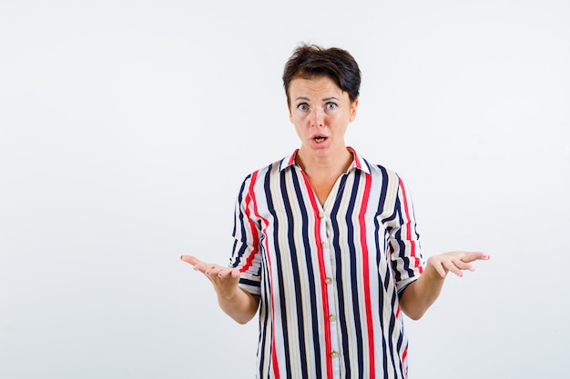 Femme mature en gardant les mains en geste perplexe en chemise rayée, vue de face.