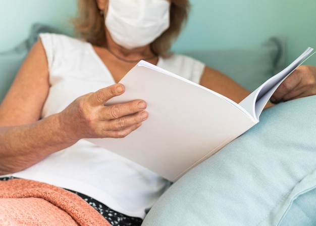 Femme mature avec masque médical à la maison pendant la pandémie en lisant un livre
